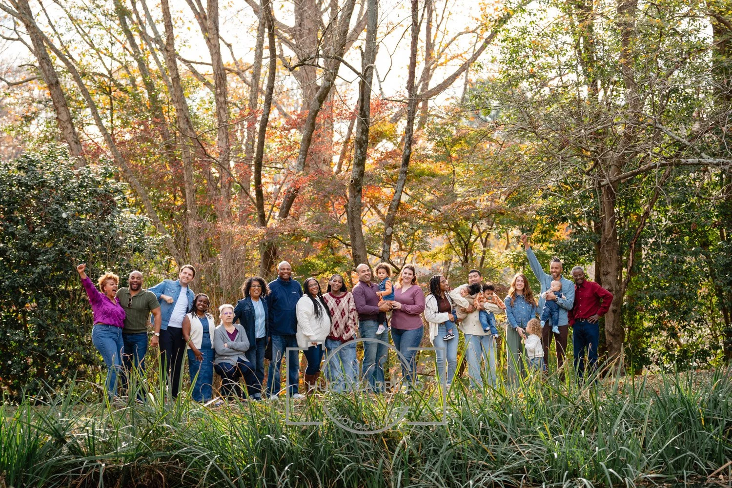 A diverse group of family and friends standing outdoors in a wooded park during fall, smiling and posing for a photo.