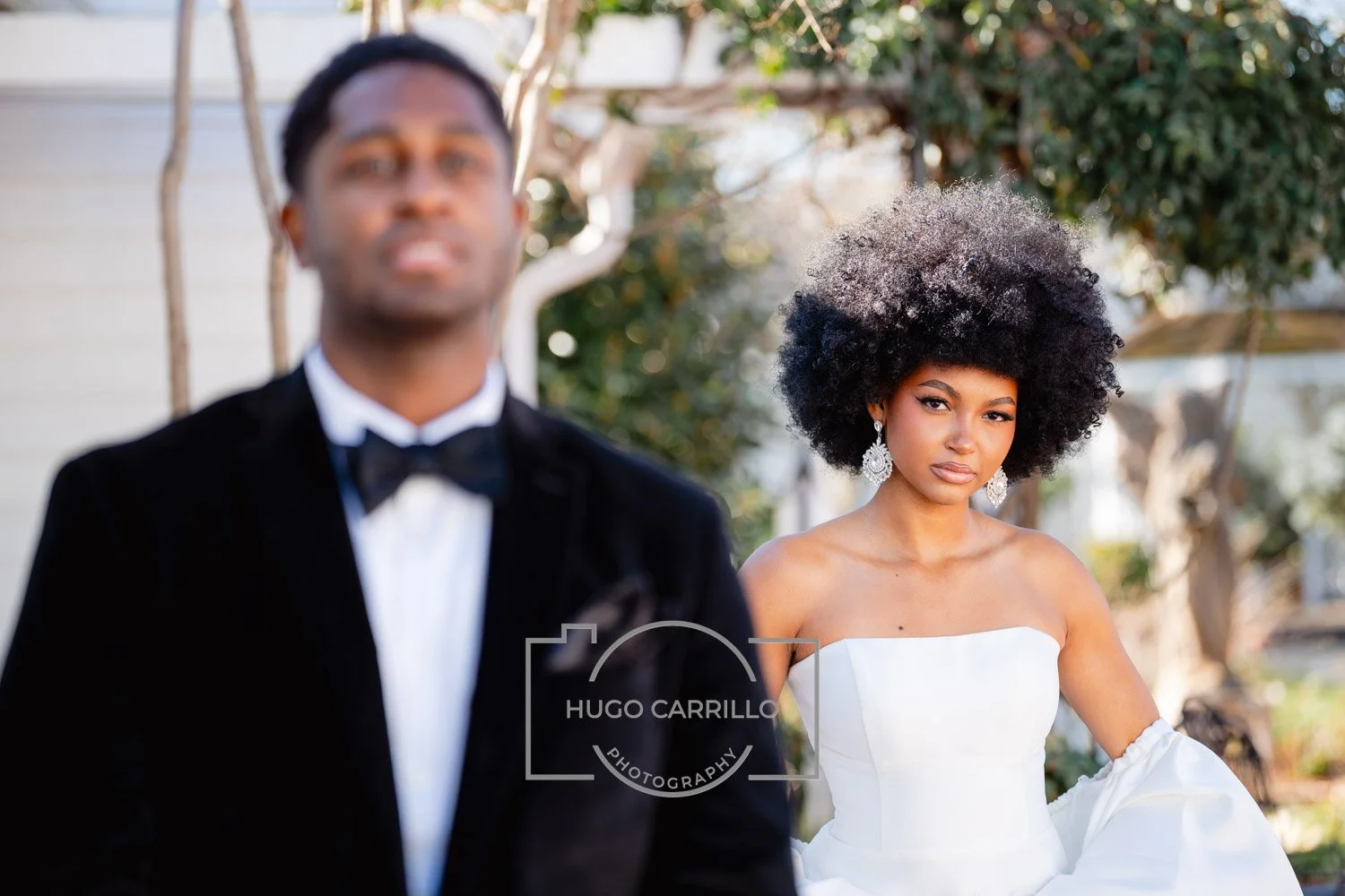 A bride with an elaborate curly afro hairstyle wearing a white strapless wedding gown and large earrings stands outdoors, while a groom in a black tuxedo with a bowtie stands slightly out of focus in the foreground.