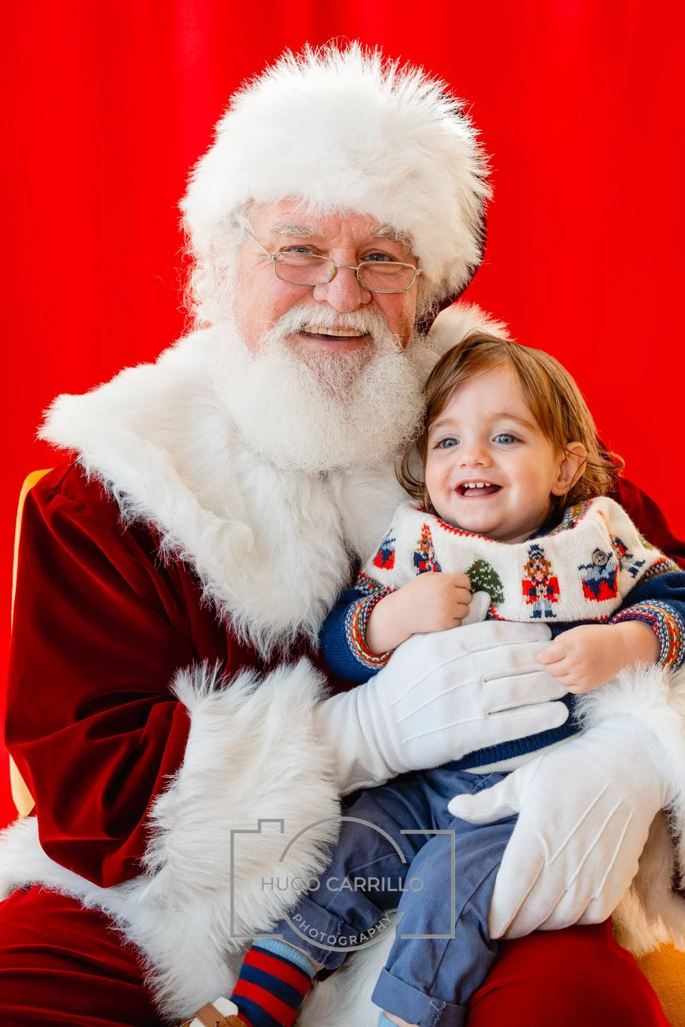 Santa Claus with a child on his lap, smiling, against a red background. Santa wears a red suit with white fur trim and glasses. The child has light brown hair, blue eyes, and is wearing a festive sweater with Christmas motifs.