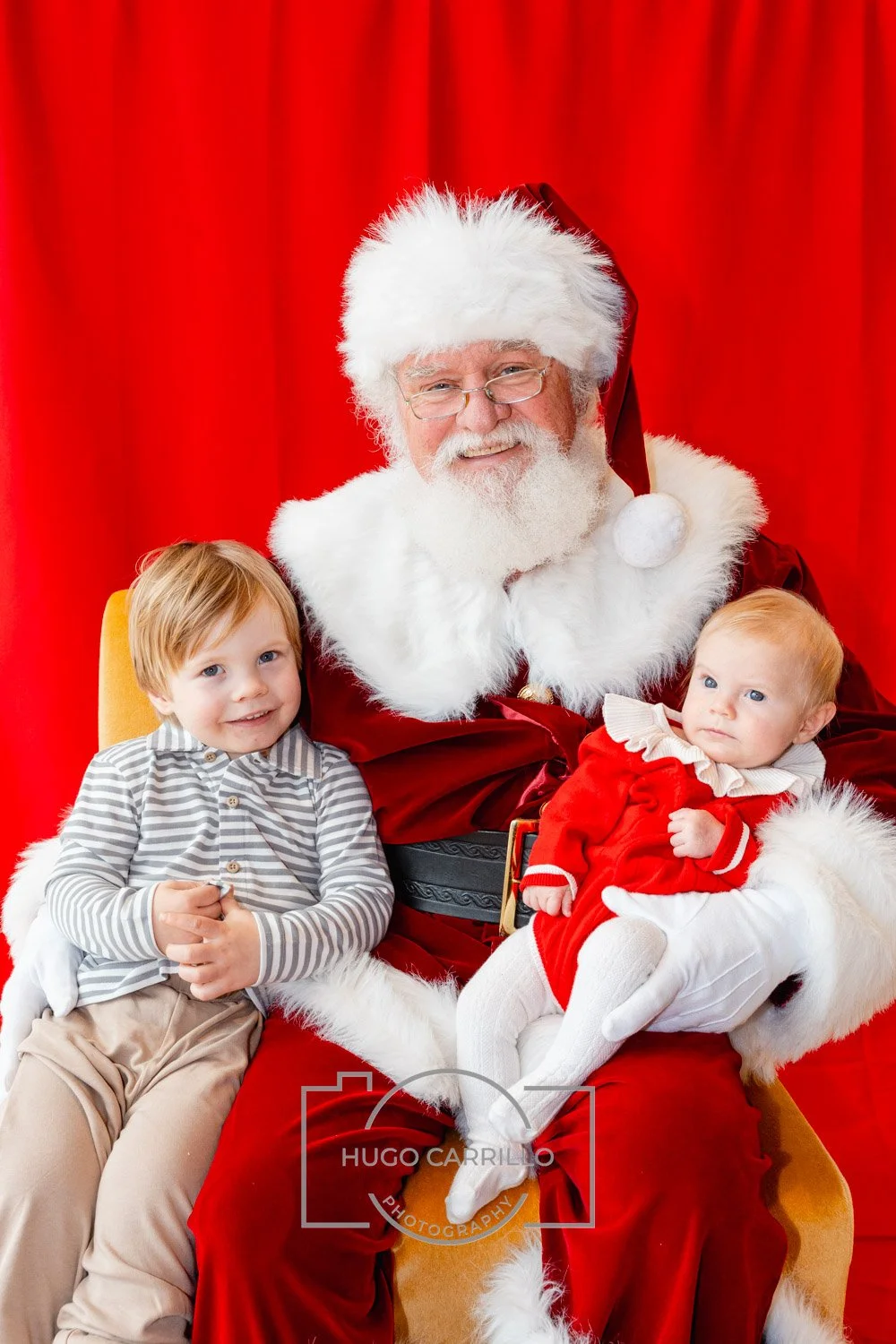 Santa Claus sitting with two young children in front of a red curtain. Santa is smiling, wearing a red suit with white fur trim and a red hat. One child, a boy with light brown hair wearing a gray and white striped shirt and beige pants, is sitting o