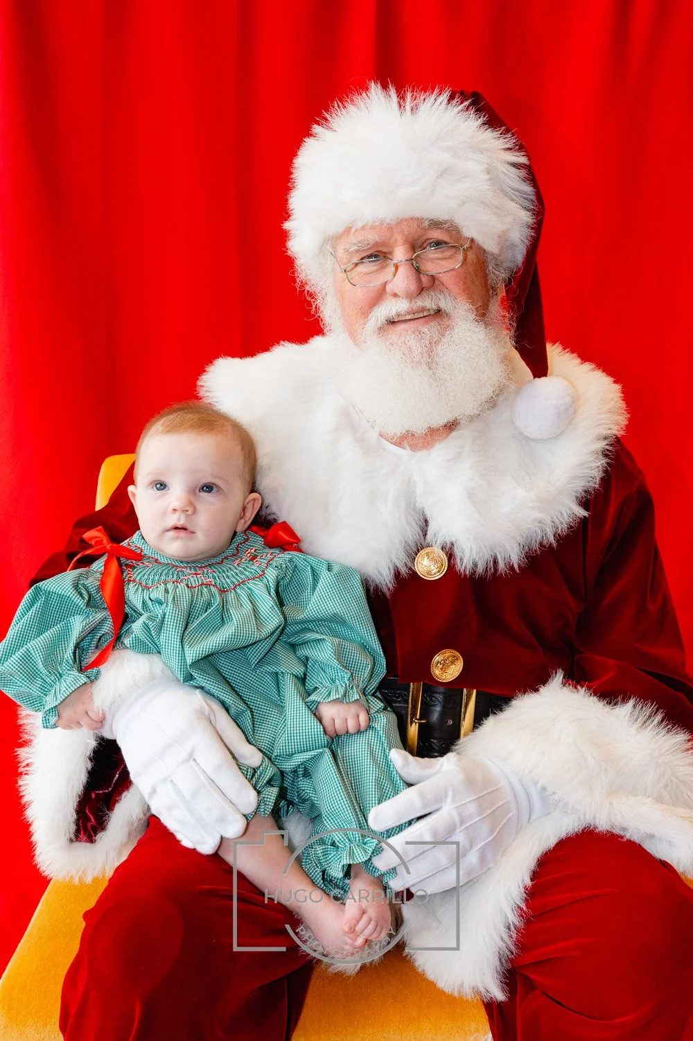 Santa Claus with a white beard, glasses, red suit with white fur trim, sitting on a yellow chair, holding a baby girl dressed in a green dress with red ribbons, against a red background.
