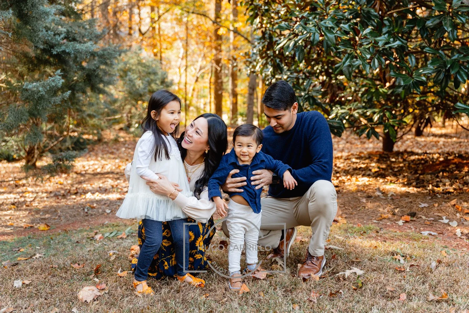 A family of four, including a mother, father, daughter, and son, enjoying fall outdoors in a wooded area with orange and green leaves, smiling and playing together.