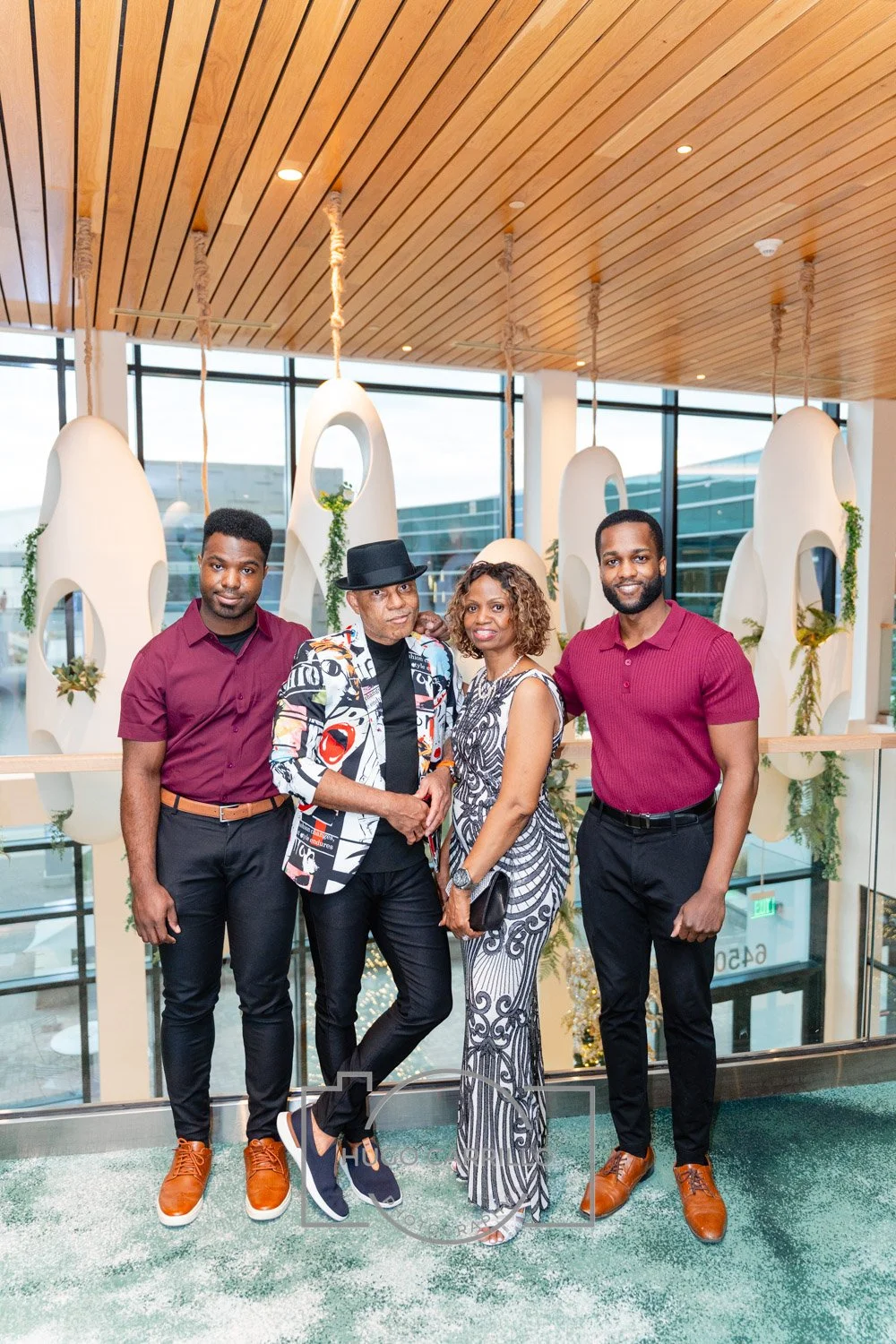 A group of four people, two men and two women, standing together inside a modern building next to a glass railing. The man on the left is wearing a maroon shirt and black pants, the man in the center is wearing a colorful patterned blazer, black hat,