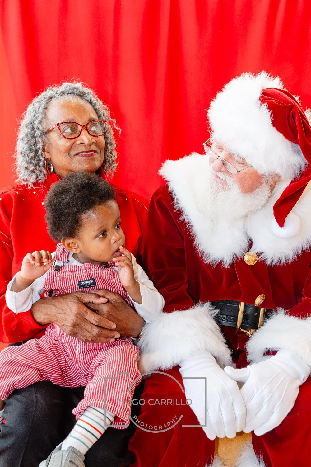 A woman, a child, and Santa Claus sitting together in front of red curtains. The woman is smiling, the child appears curious, and Santa is looking at the woman.