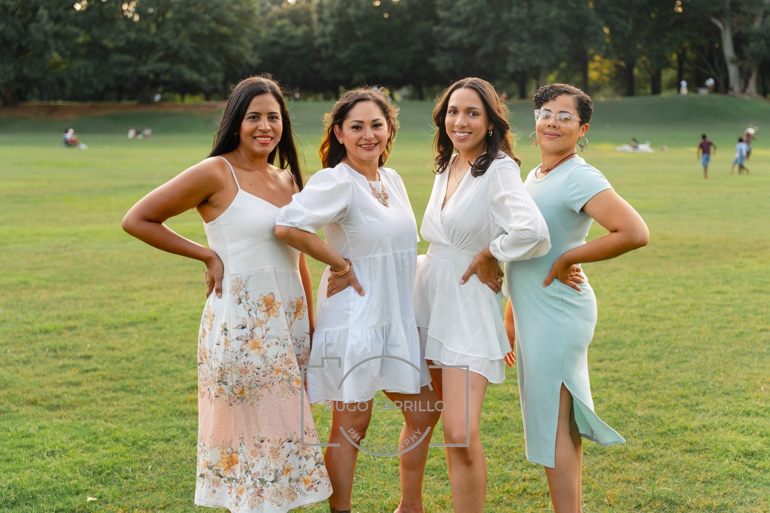 Four women standing on a grassy park, smiling at the camera, dressed in light-colored dresses, with trees in the background.