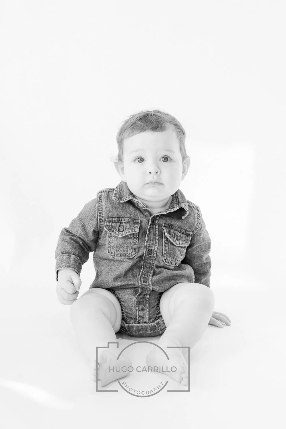 Black and white photo of a baby sitting on the floor, wearing a denim jacket and shorts, with a worried expression on their face.