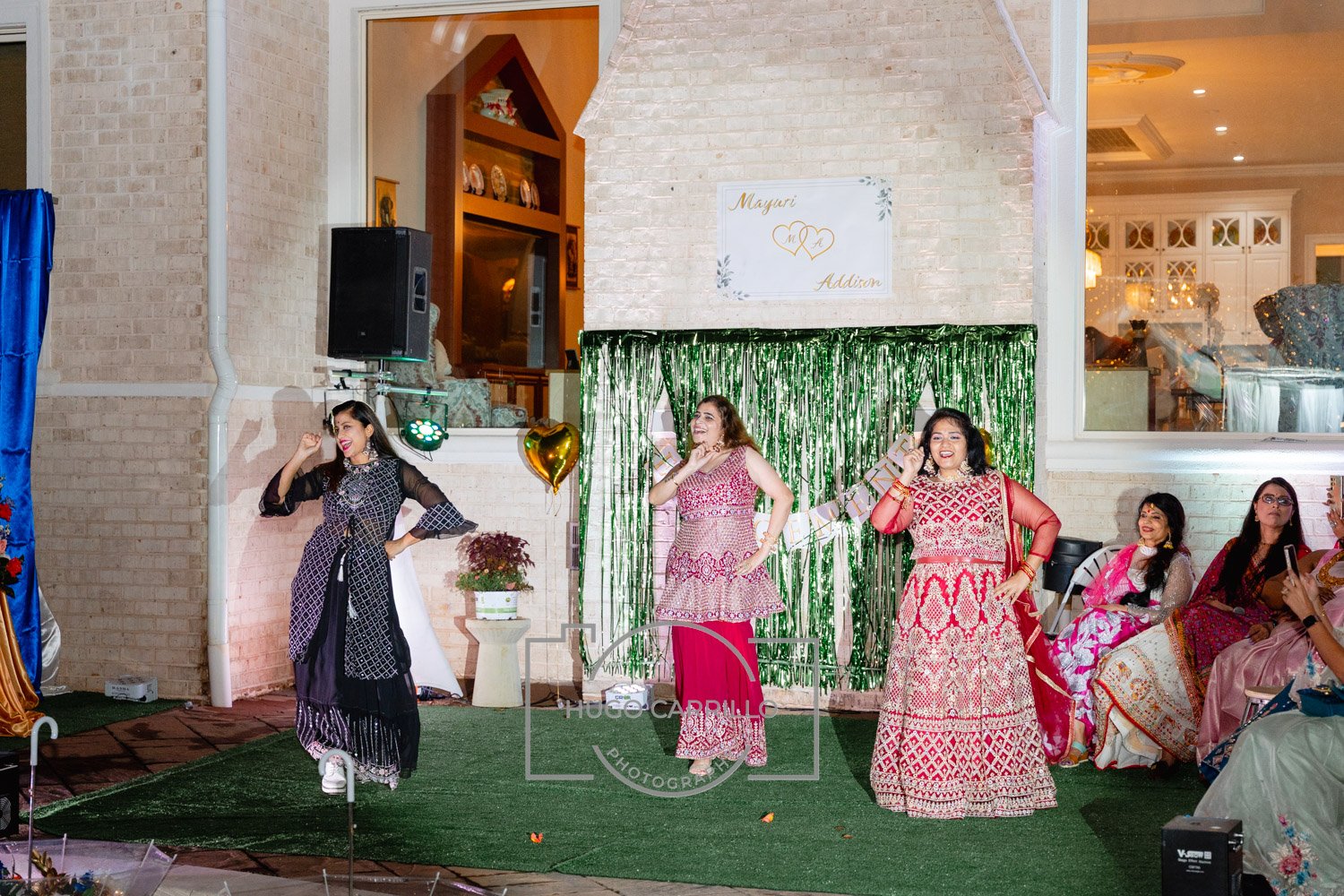 Women dressed in traditional South Asian attire performing a dance on a green grass-like stage at a celebration, with some sitting and watching and others taking photos, including a decorative backdrop with green streamers, balloons, and a sign that 