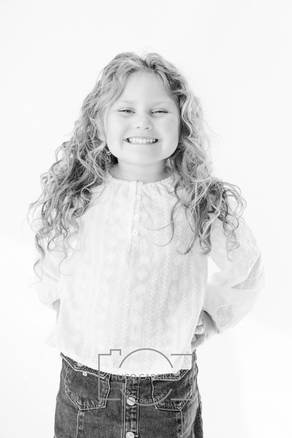 A young girl with long curly hair smiling and posing with hands on hips, wearing a light-colored embroidered blouse and denim skirt, against a plain white background.