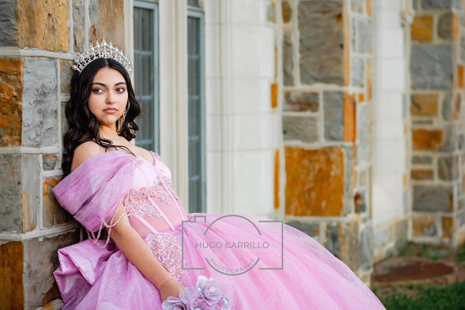 A quinceañera in a pink princess dress wearing a silver crown, sitting against a stone wall with a building and windows in the background.