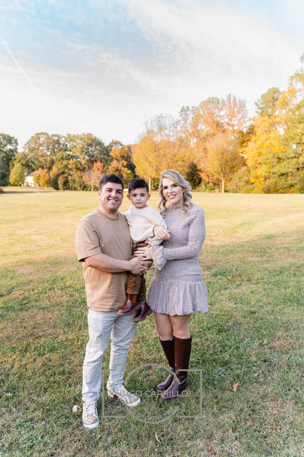 A smiling family of three standing on grass in a park during fall, with trees showing autumn colors in the background.
