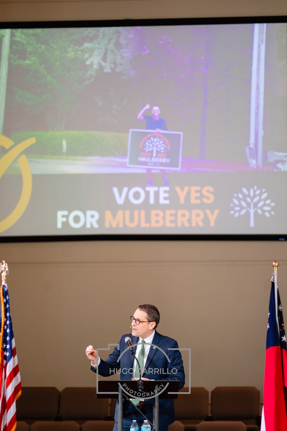 A man in a suit and glasses speaking at a podium in front of a large screen and two American flags. The screen shows a campaign advertisement with a hashtag and the text "VOTE YES FOR MULBERRY" along with an image of a person making a fist.