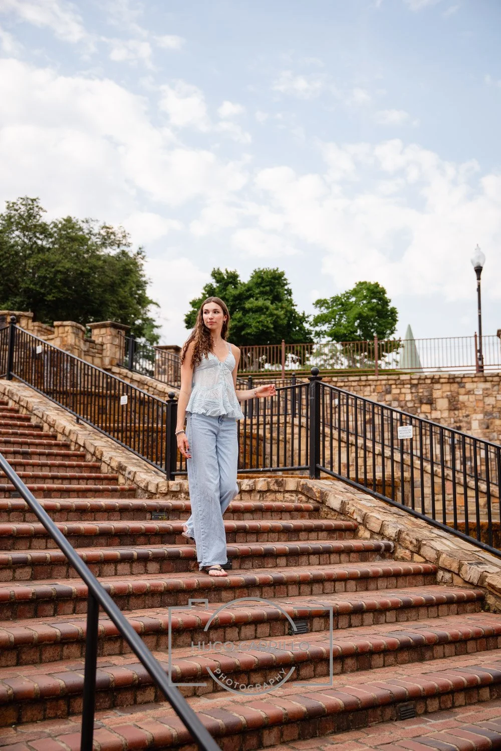 A young woman walking down an outdoor brick staircase with black metal railings, surrounded by trees and a stone wall, under a partly cloudy sky.