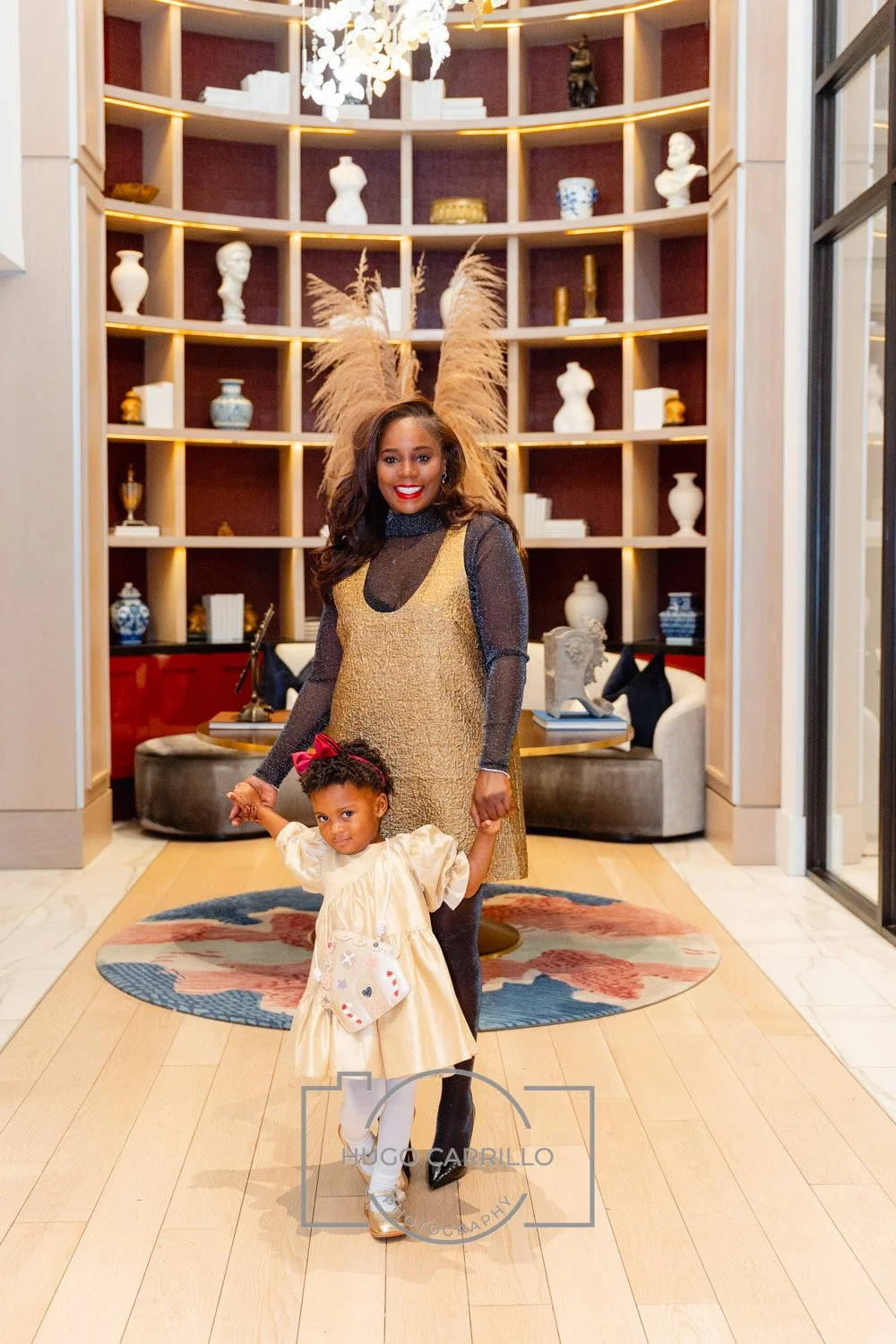 A woman and a young girl holding hands in a modern, elegantly decorated lobby with wooden shelves filled with various decorative vases and sculptures. The woman is dressed in a gold and black outfit, and the girl wears a cream-colored dress with a re