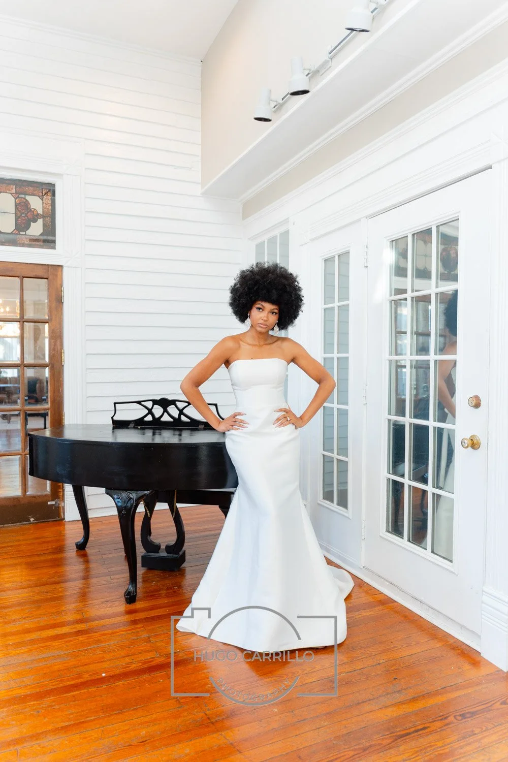 A woman in a strapless white wedding gown standing indoors near a black grand piano, with white paneled walls and glass-paneled doors behind her.