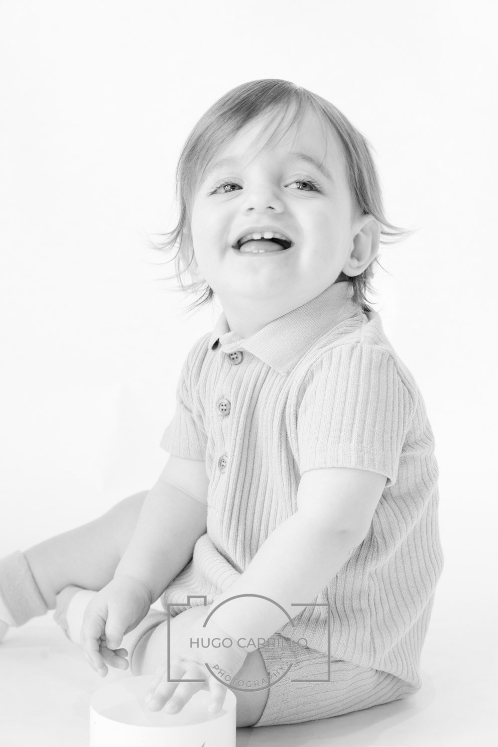 Black and white photo of a smiling young child with light hair, wearing a striped polo shirt, sitting on the floor with legs crossed.