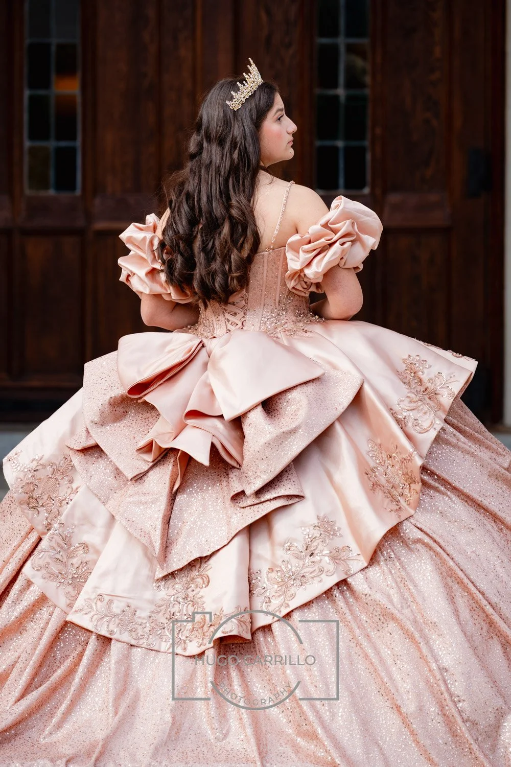 A quinceañera in an elegant pink ball gown with ruffles and embroidery, wearing a tiara, sitting in front of a wooden background.