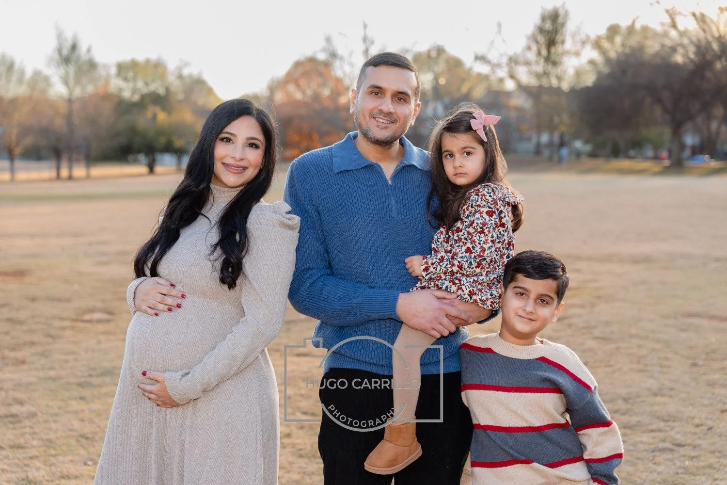 A family of four standing outdoors in a park during autumn. The pregnant woman is on the left, wearing a beige sweater dress, and smiling. The man in the center is holding a young girl with dark hair and a pink bow, wearing a floral dress. A young bo