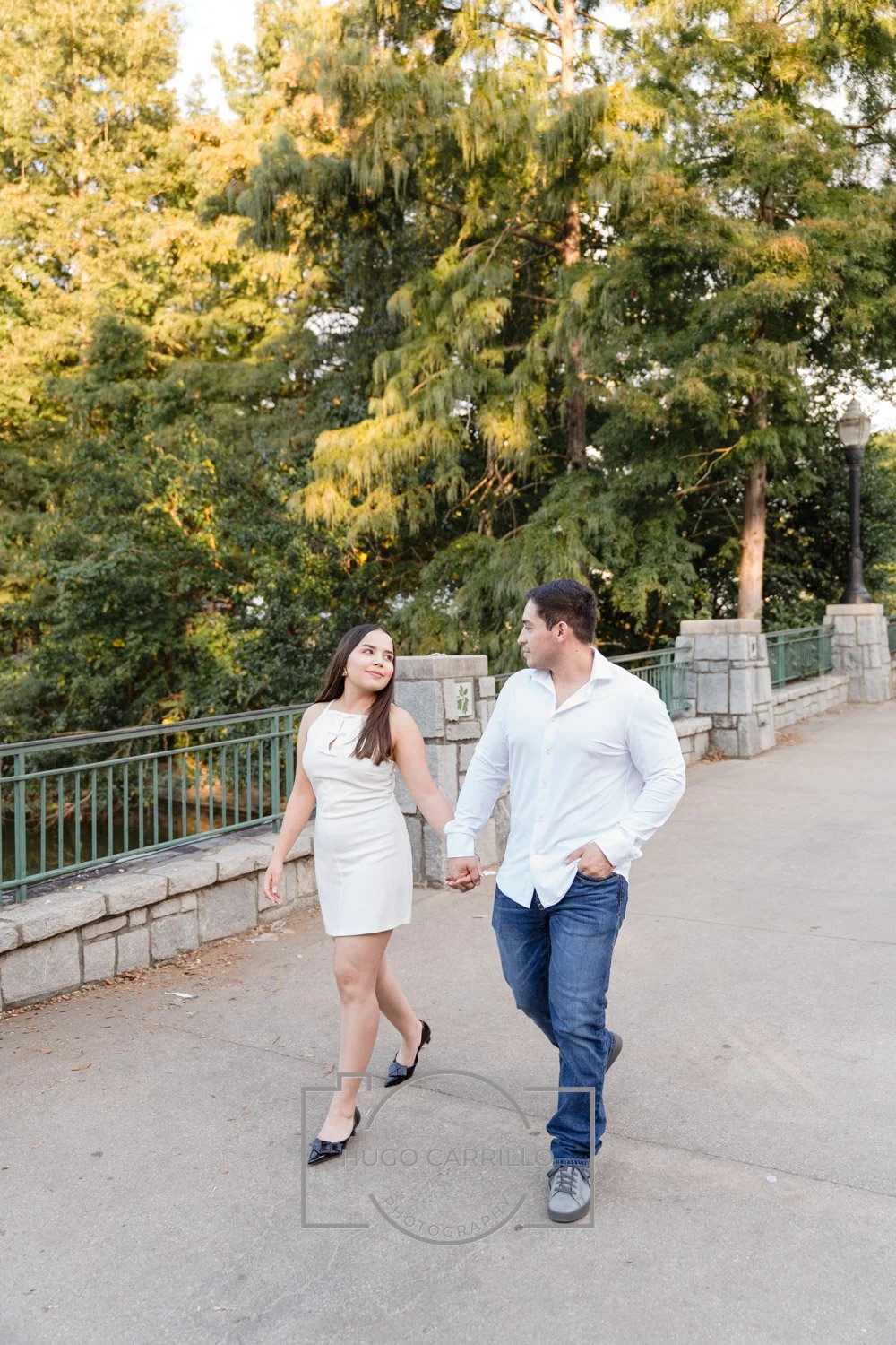 A couple holding hands and walking along a park path surrounded by tall green trees, with a stone and metal railing and a lamppost in the background.
