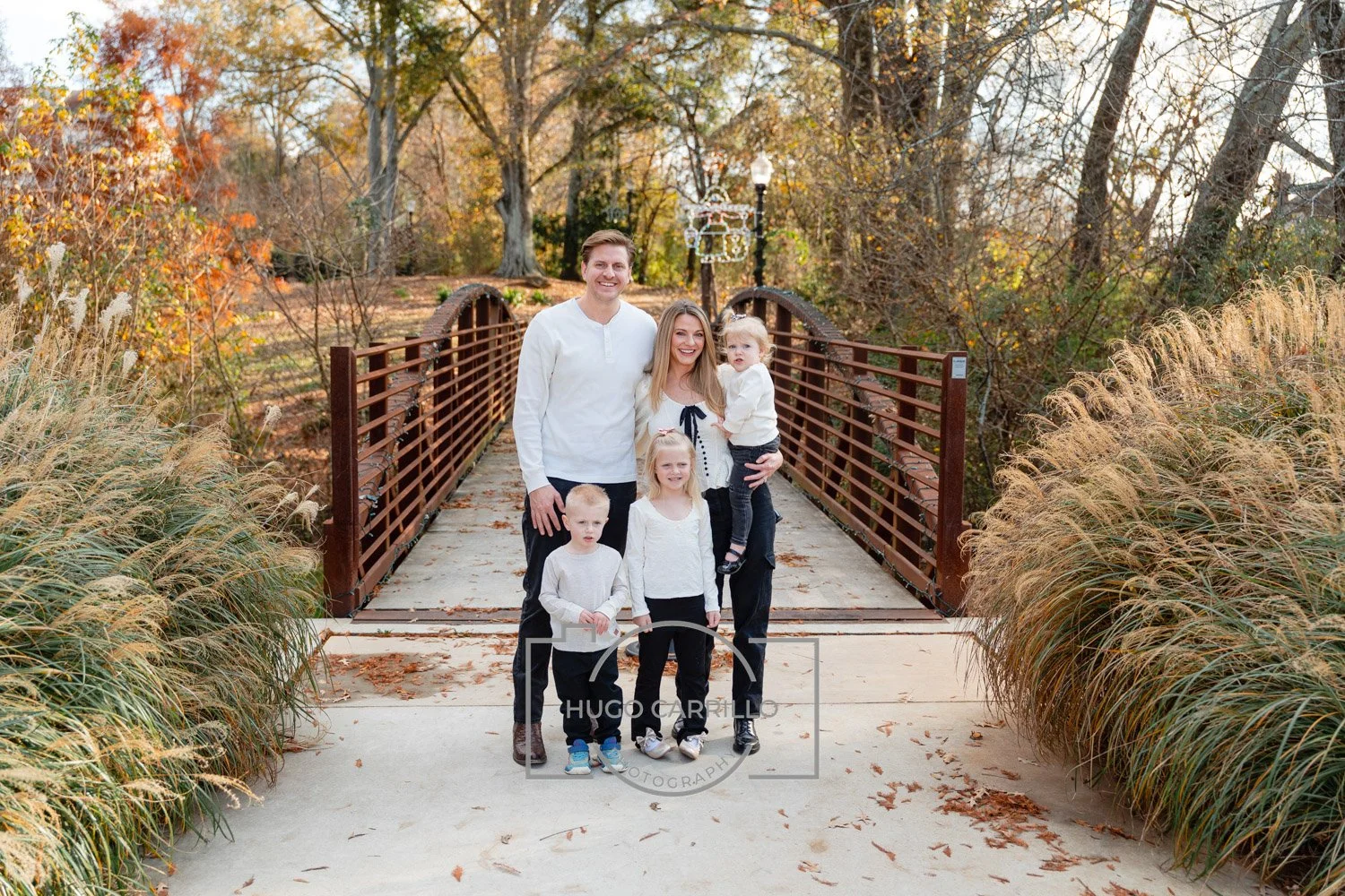 A family of six posing on a small wooden bridge in a park with autumn foliage, including trees with orange, yellow, and green leaves, and tall grasses on either side.