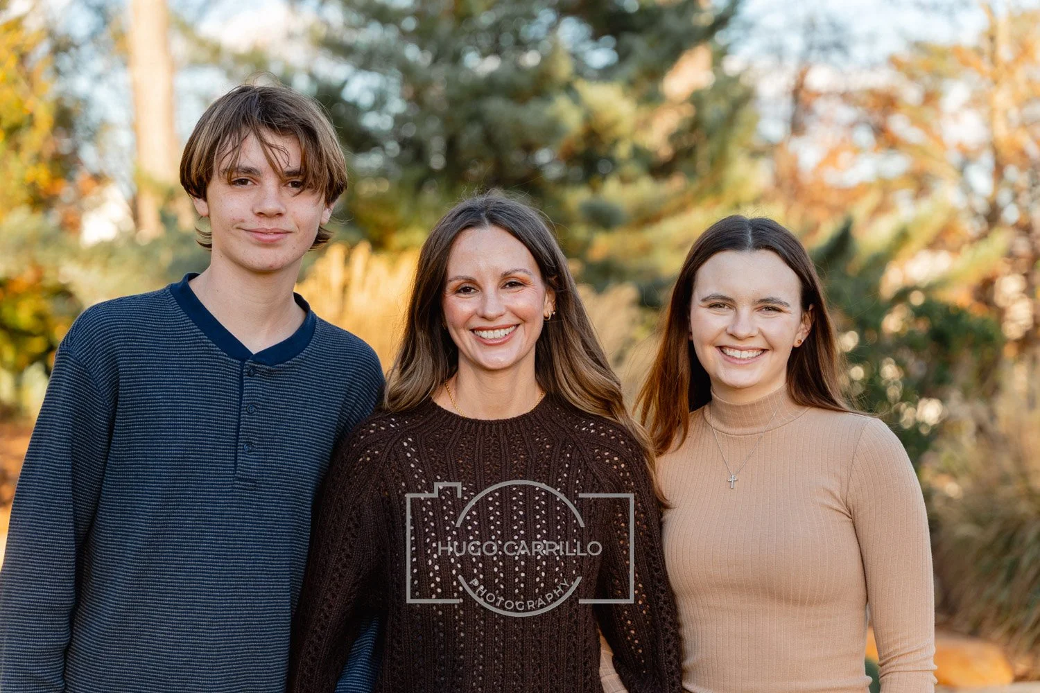 A woman and two teenagers smiling outdoors in fall with trees and foliage in the background.