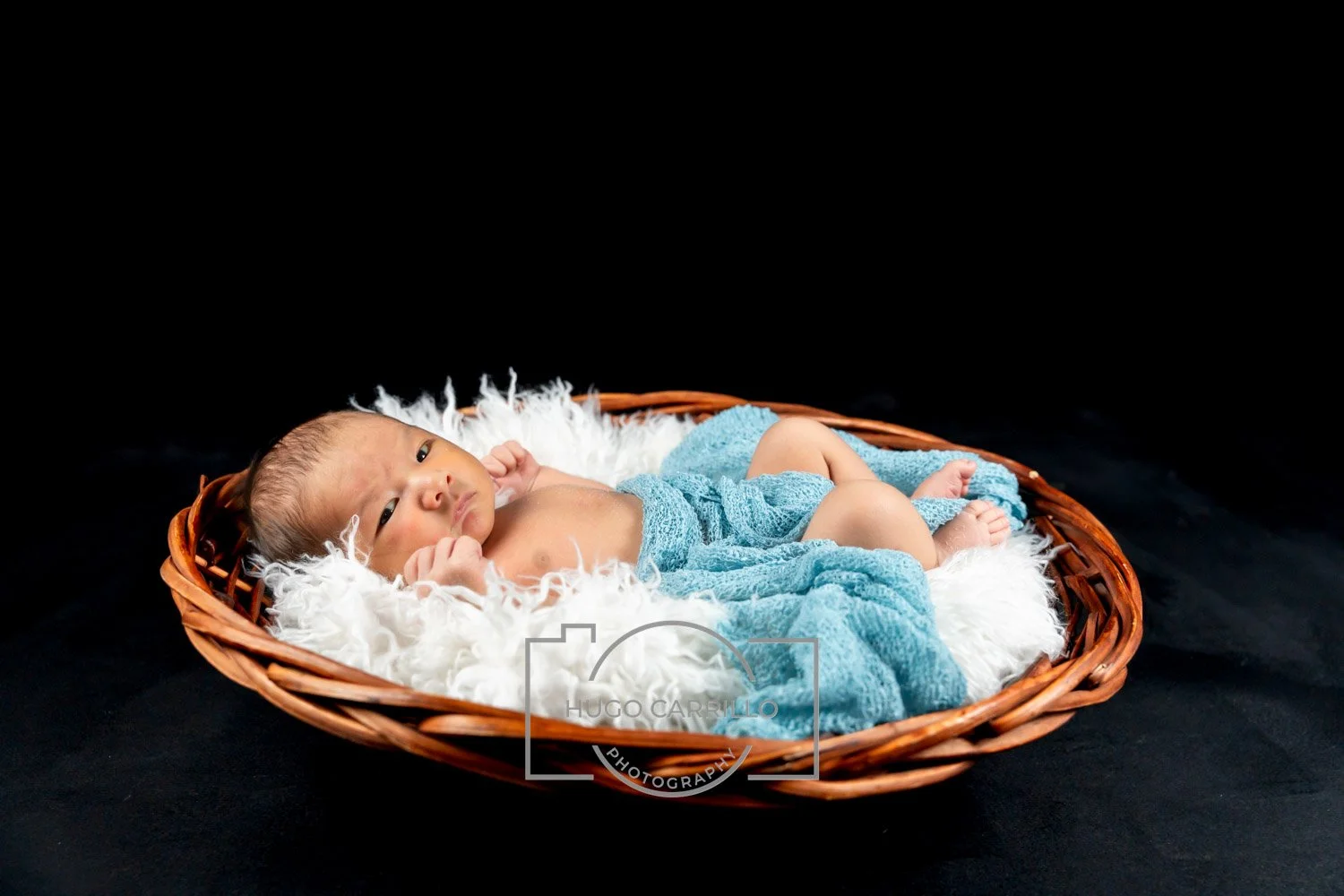 Newborn baby lying in a wicker basket on white bedding against a black background, wrapped in a blue blanket.
