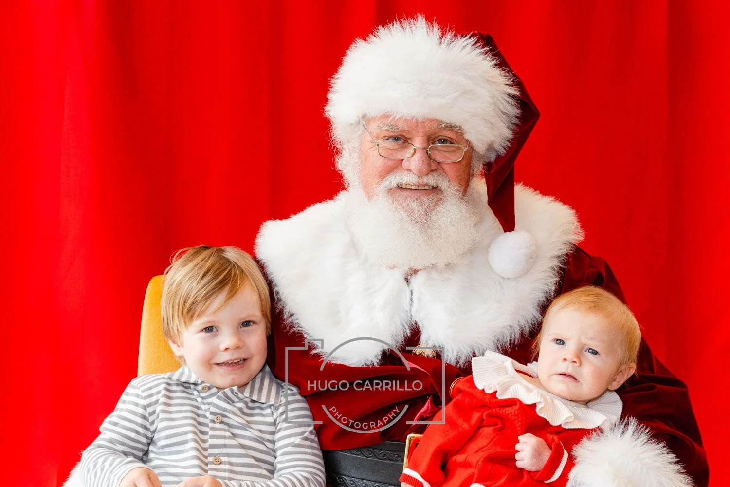 Santa Claus with two young children in front of a red curtain, one child smiling and the other looking confused.