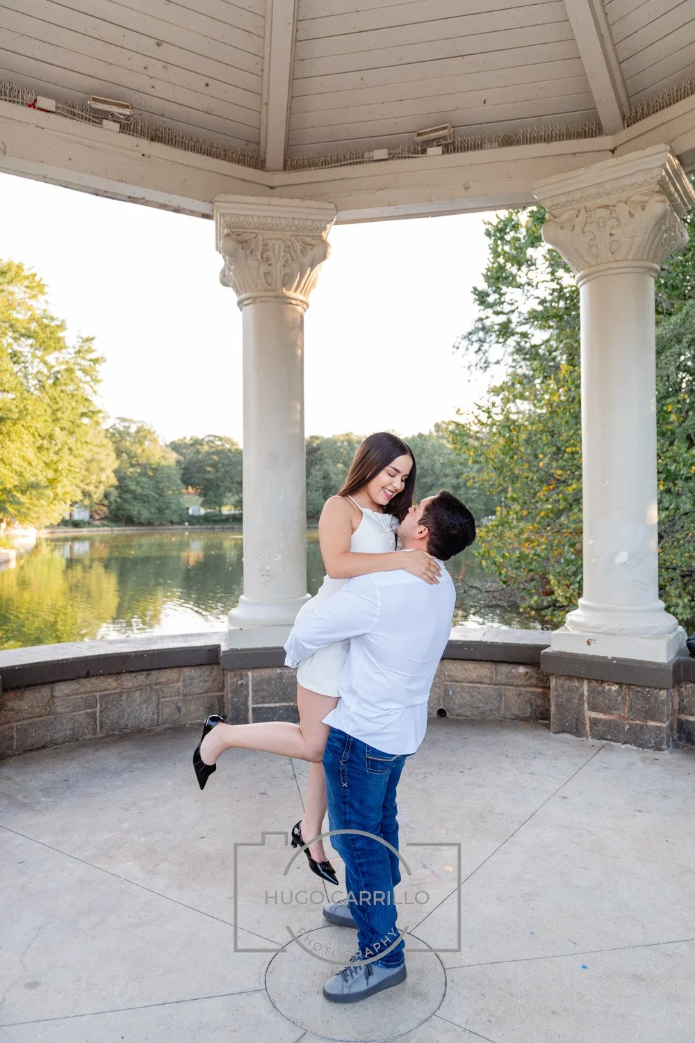 A couple in white clothing, with the man holding the woman in his arms, smiling and looking at each other, standing under a decorative pavilion near a body of water with trees in the background.