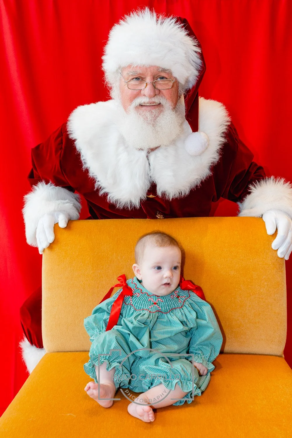Santa Claus dressed in a red velvet suit with white fur trim, holding a yellow chair, with a baby girl sitting on it, against a red curtain background.