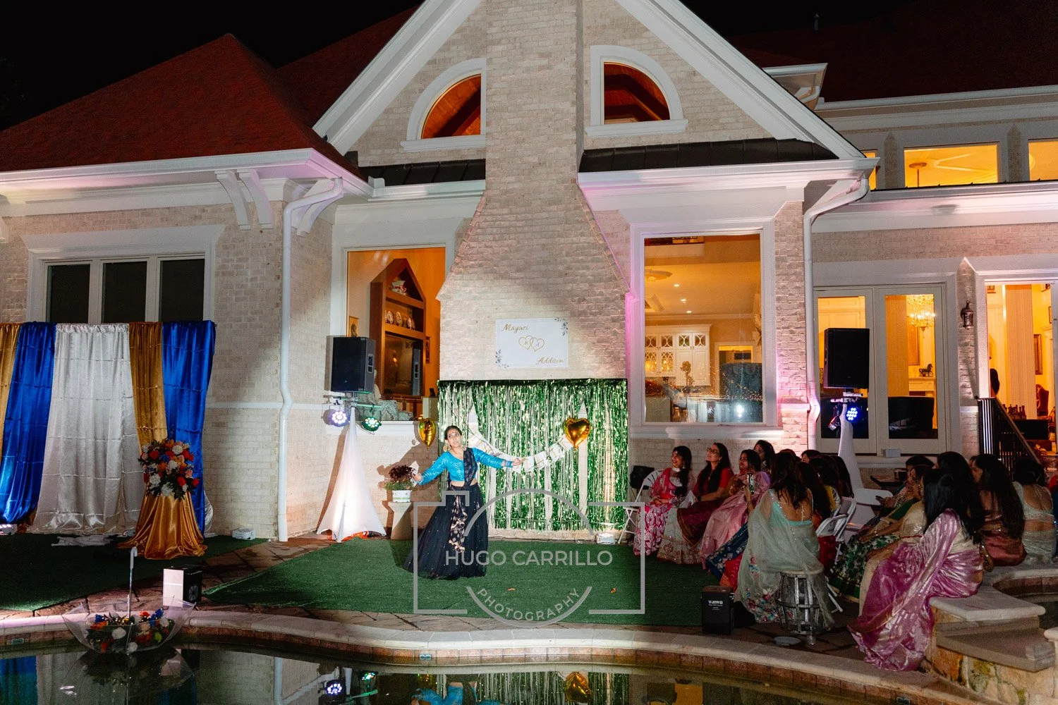 A cultural celebration or party at a backyard or patio of a house at night, where a woman in traditional attire performs in front of an audience, with decorations including gold heart-shaped balloons, colorful curtains, and a whiteboard with initials