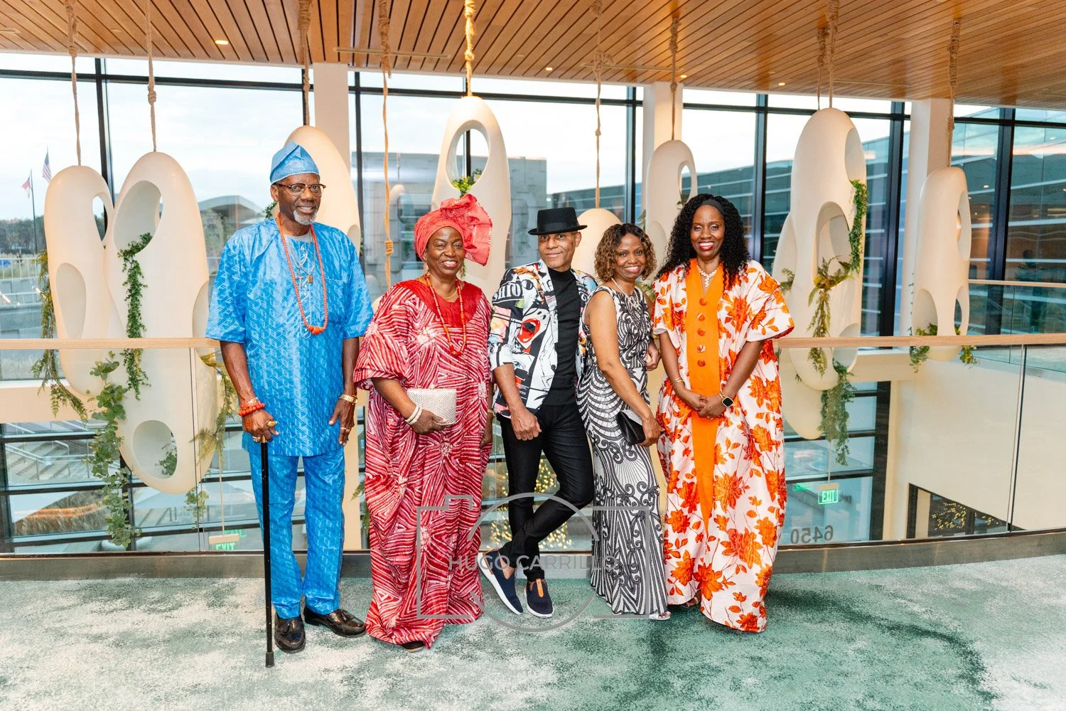 Six people standing together in a modern indoor space, dressed in colorful, traditional African-inspired clothing, with large decorative white elements and glass windows in the background.