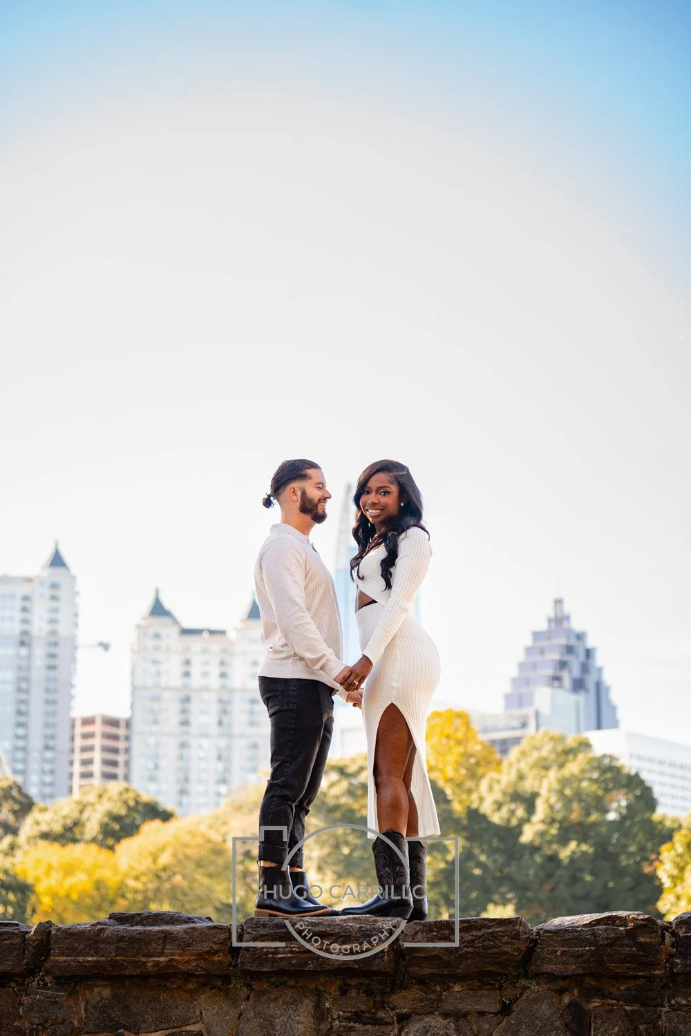 A couple holding hands and smiling in a city park with tall buildings and trees in the background during daytime.
