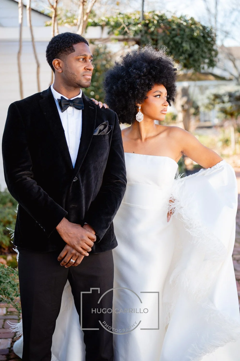 A man and woman dressed in formal wedding attire standing outdoors, with trees and a house in the background.