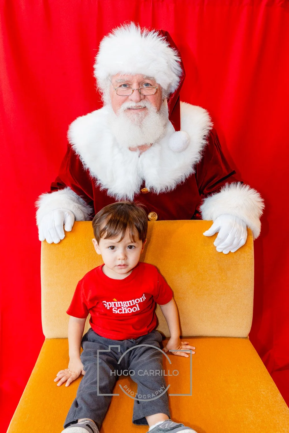 A young boy sitting on an orange chair, looking confused or upset, with Santa Claus standing behind him. Santa is dressed in a traditional red and white suit with glasses, white gloves, and a white beard, and is smiling. The background is red.