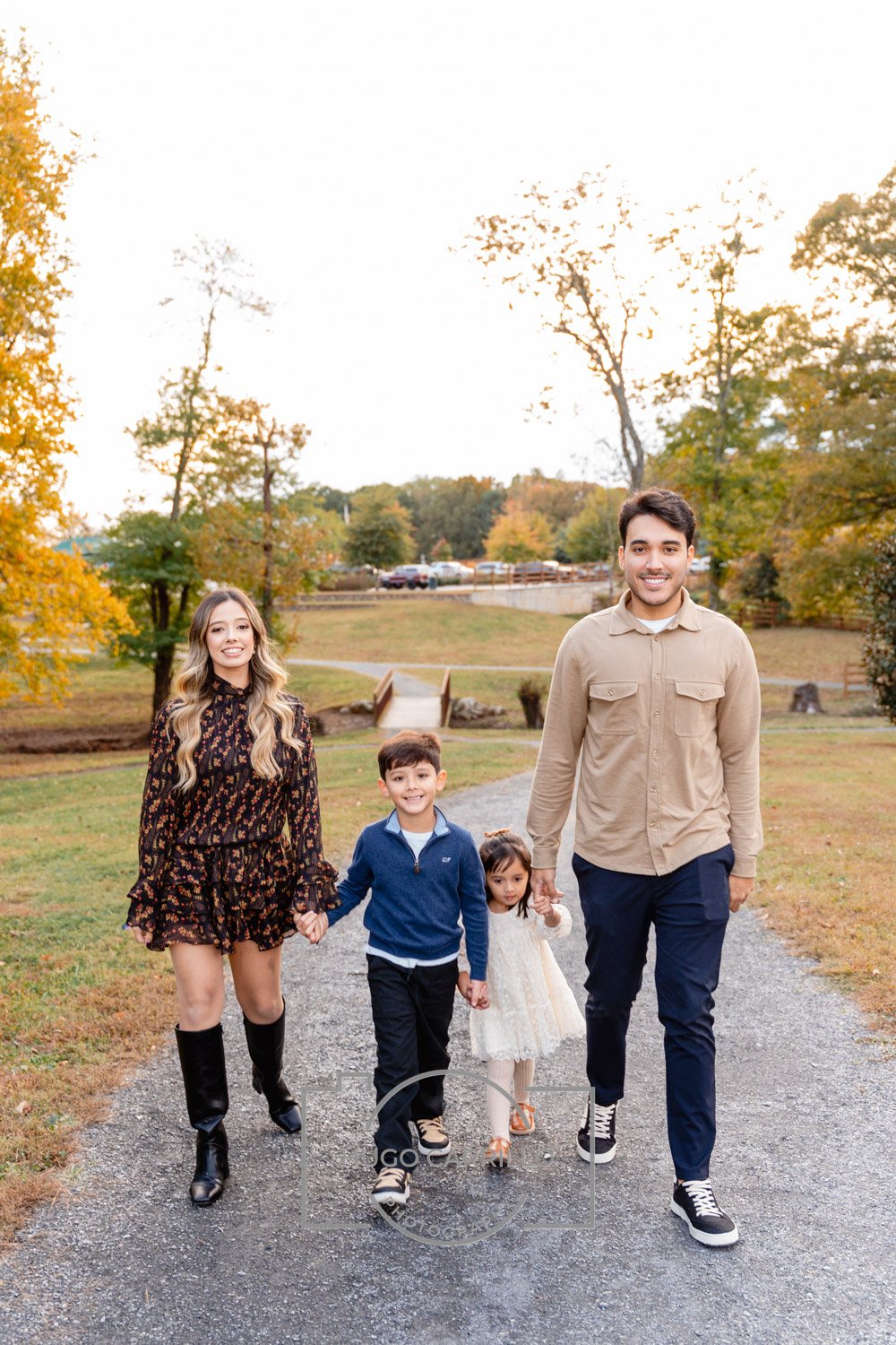 A family of four walking in a park during autumn, holding hands and smiling. The family includes a woman with long blonde hair wearing a dark floral dress and black boots, a man with short dark hair in a beige shirt and navy pants, a young boy in a b