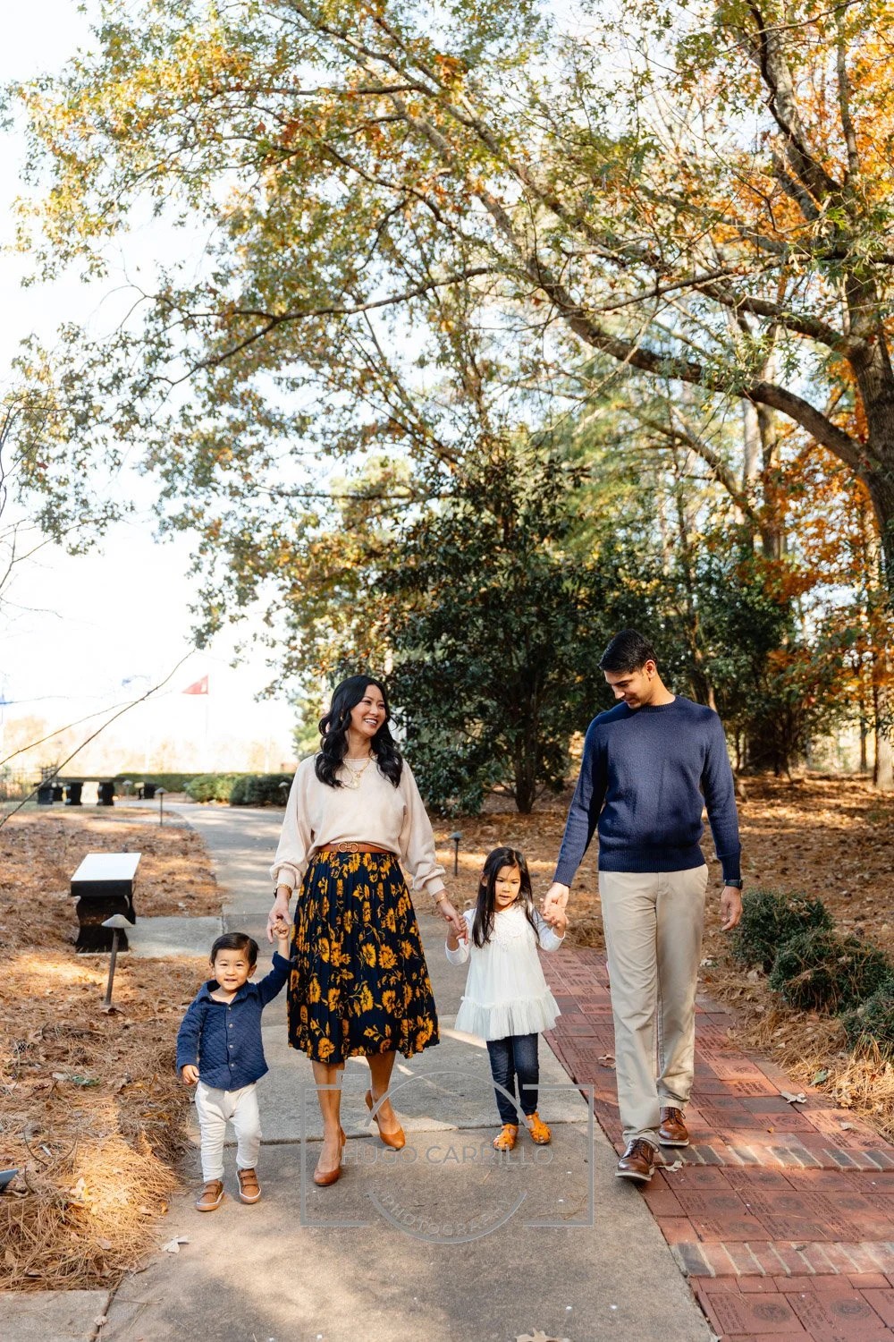 Family walking outdoors on a fall day, holding hands and smiling, surrounded by autumn trees and fallen leaves.