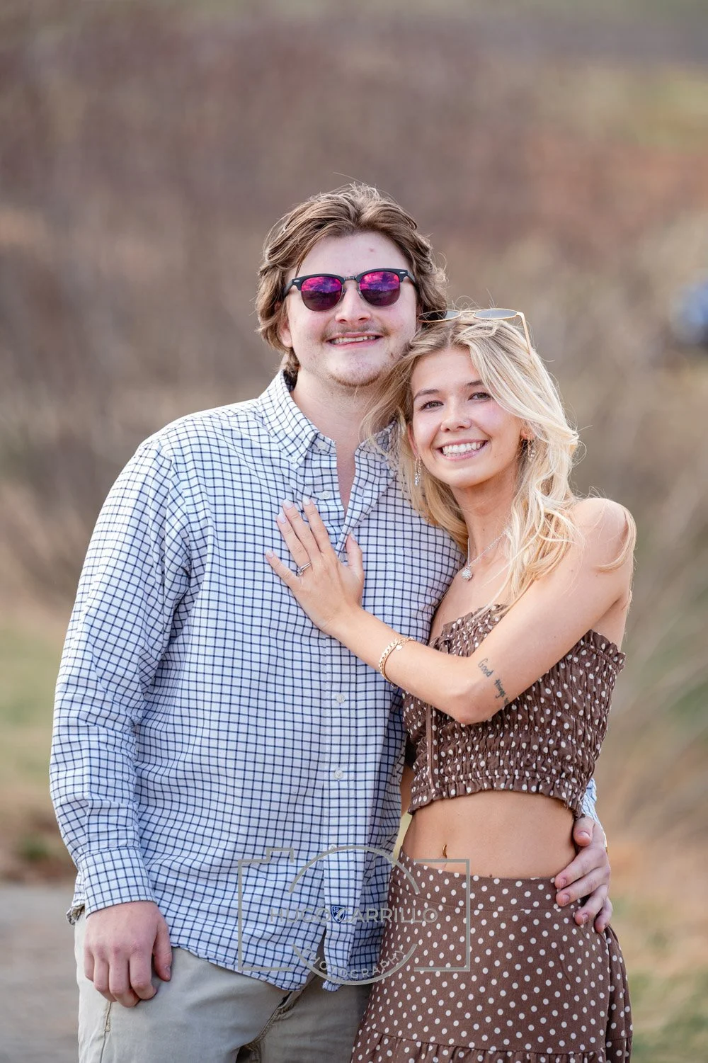 A young couple smiling and embracing outdoors, with a blurred natural background. The man has wavy hair, sunglasses, and a checkered shirt. The woman has long, wavy blonde hair, a wide smile, and is wearing a polka dot crop top and skirt.