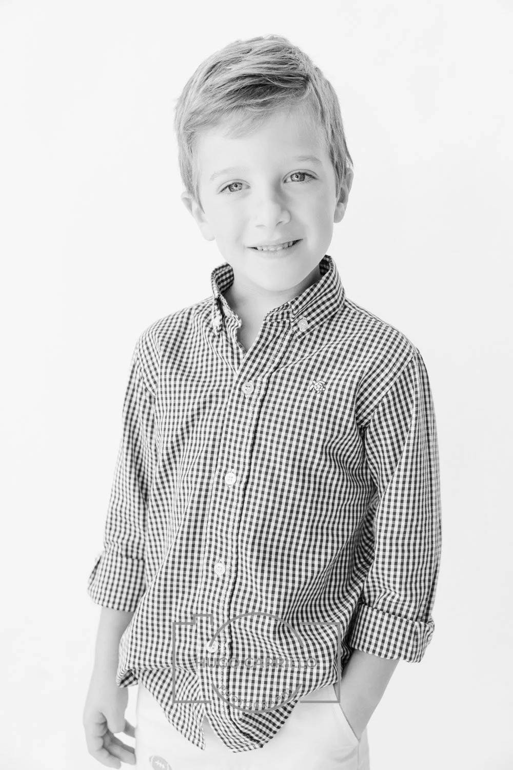 Black and white portrait of a young boy with light hair, smiling, wearing a checkered button-up shirt with sleeves rolled up, standing against a plain background.