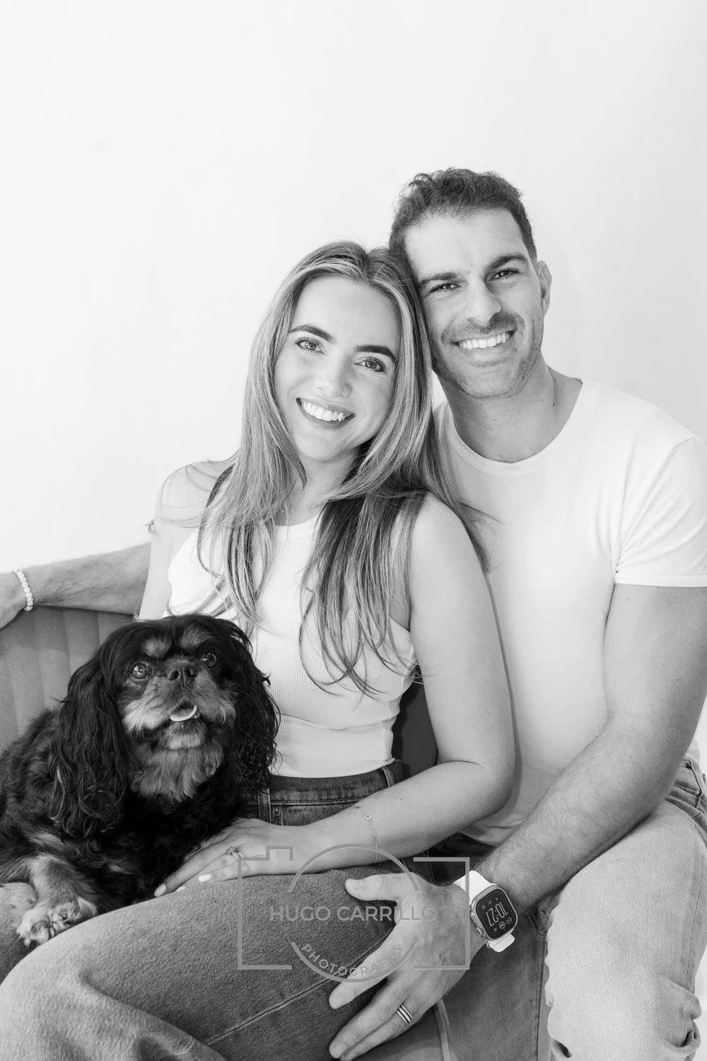 Black and white photo of a smiling woman and man sitting close together, with a small dog in front of them. The woman has long hair and the man has short hair. They are both wearing casual clothing, and the woman is wearing a bracelet.
