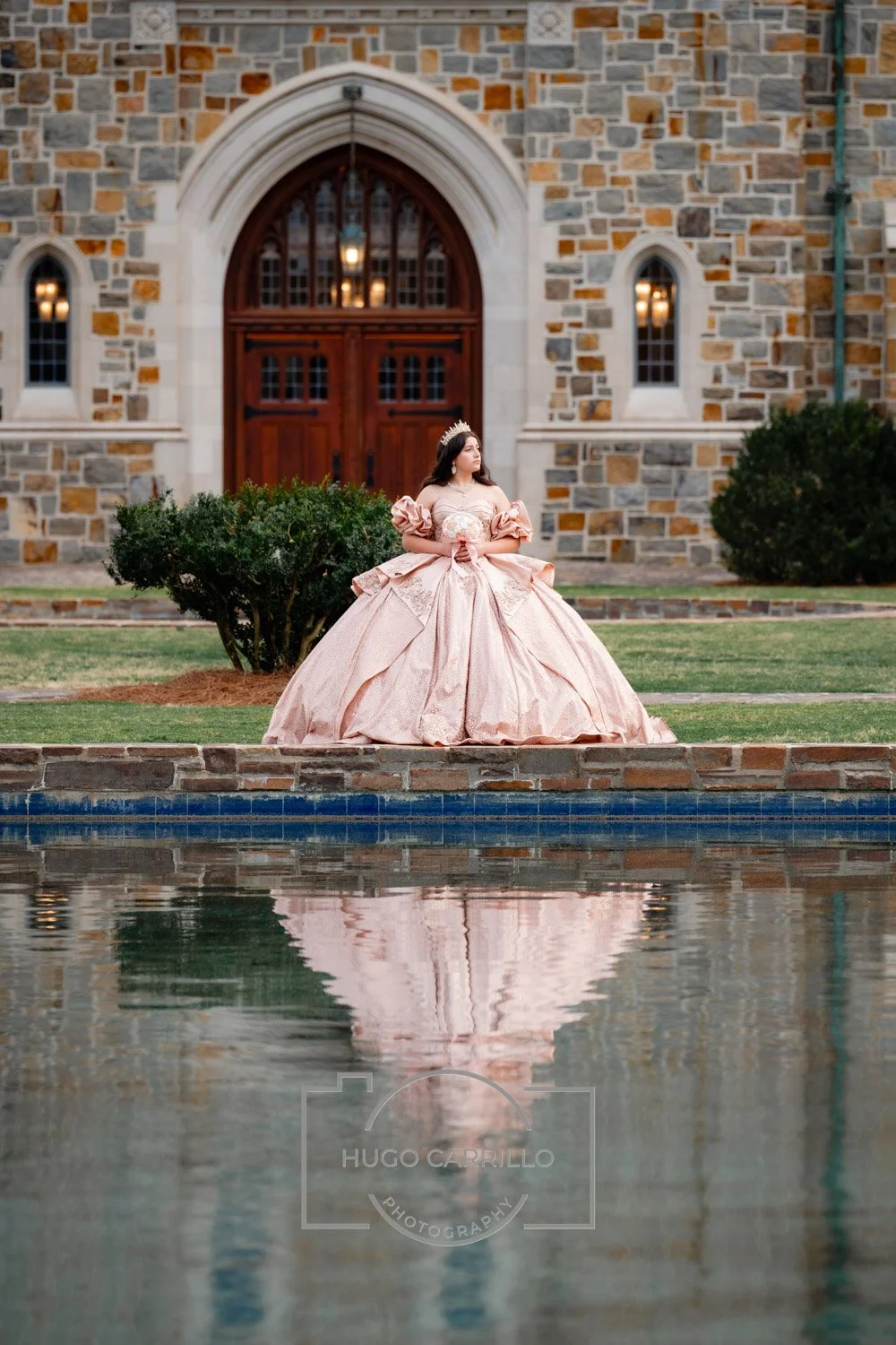 A quinceañera in a pink, royal-looking gown with puffed sleeves and a large skirt, wearing a tiara, stands outdoors in front of a castle-like building, with a pond reflecting her image.