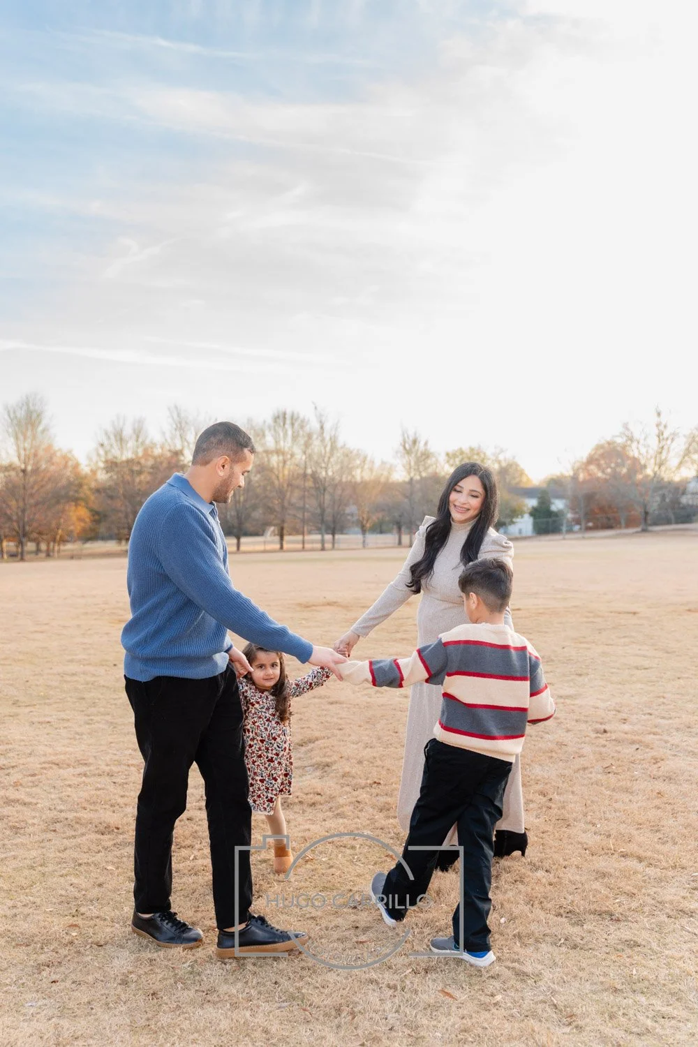 A family of four holding hands and playing together outdoors on a grassy field during autumn, with trees in the background and a cloudy sky.