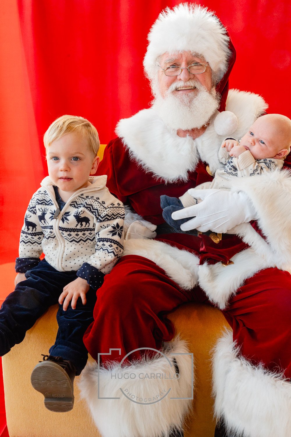 Santa Claus sitting with two young boys in front of a red background, one boy sitting on his lap and the other sitting beside him, both children wearing Christmas sweaters.
