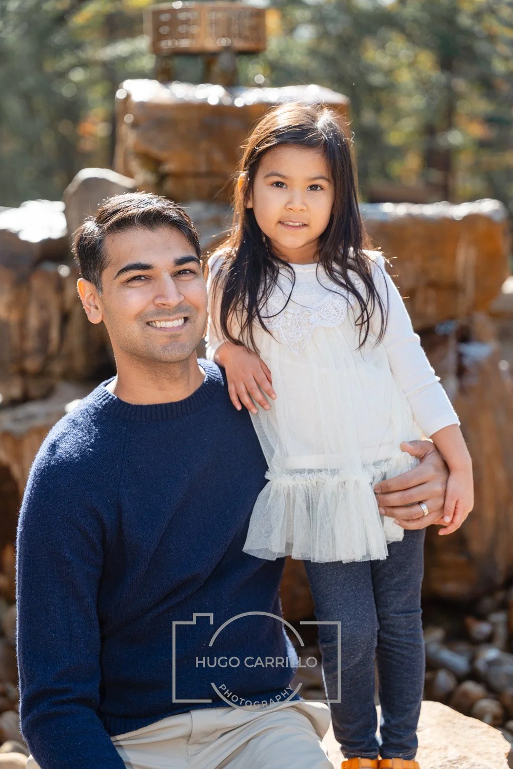 A man with dark hair, wearing a navy blue sweater and beige pants, holding a young girl with long dark hair, dressed in a white dress and gray leggings, outdoors near a waterfall with rocks and trees in the background.