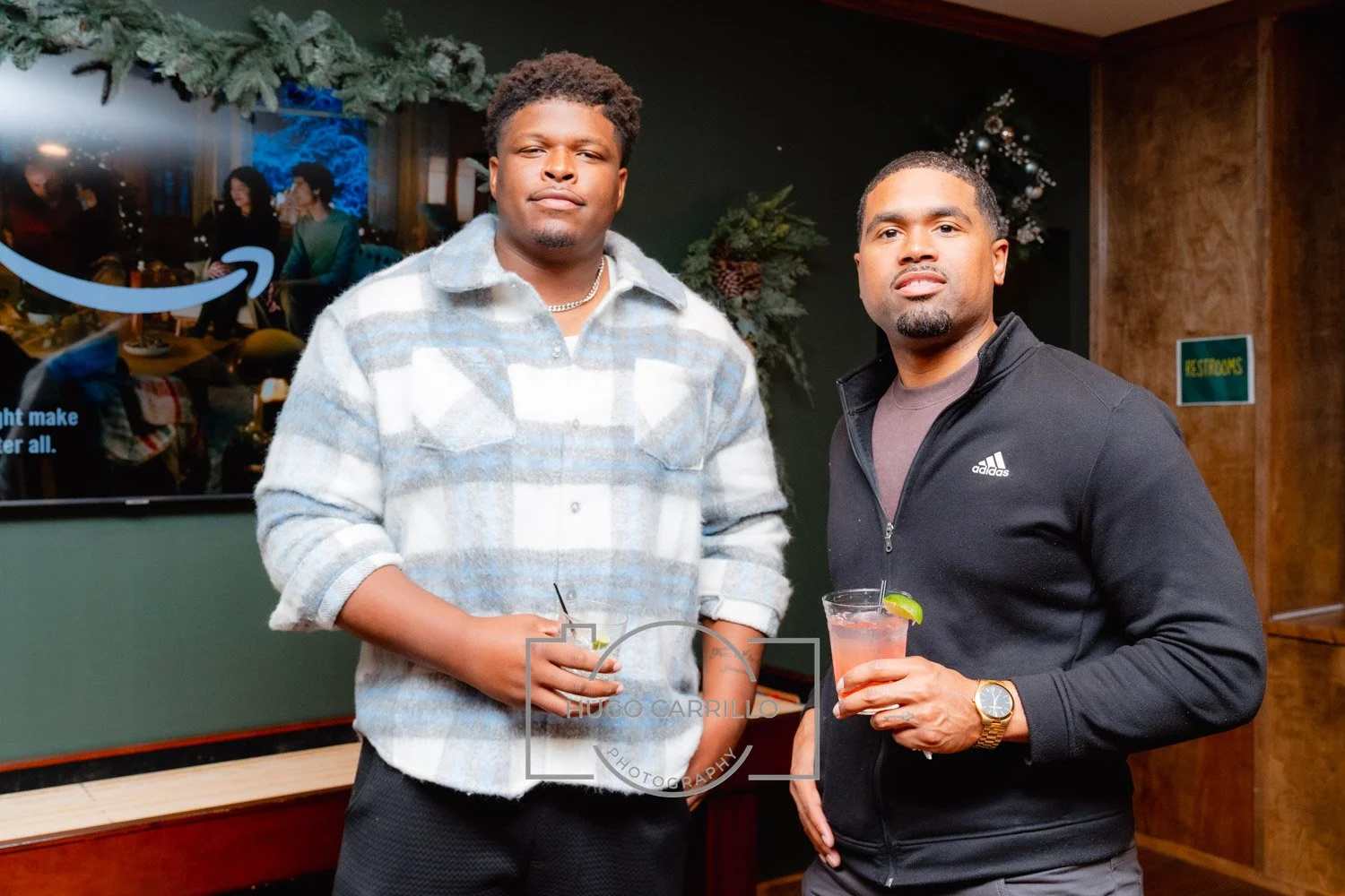 Two men with short hair, one wearing a plaid shirt and the other a black Adidas jacket, holding drinks in a bar or restaurant with holiday decorations.