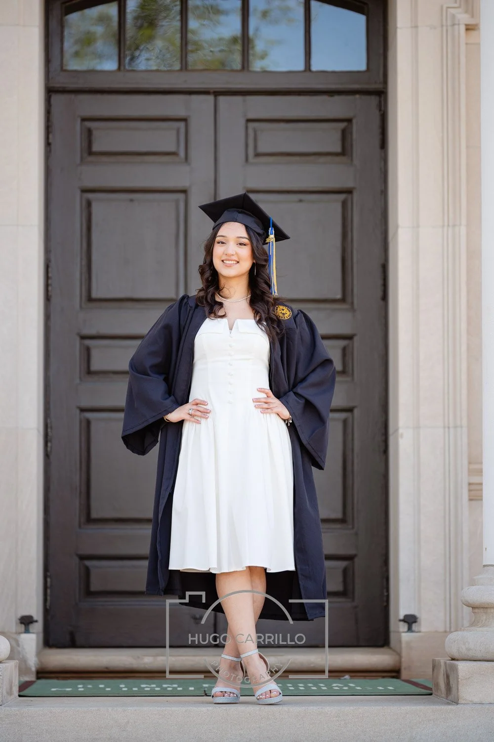 Young woman in a graduation cap and gown standing on steps in front of a large wooden door, smiling for a photo.