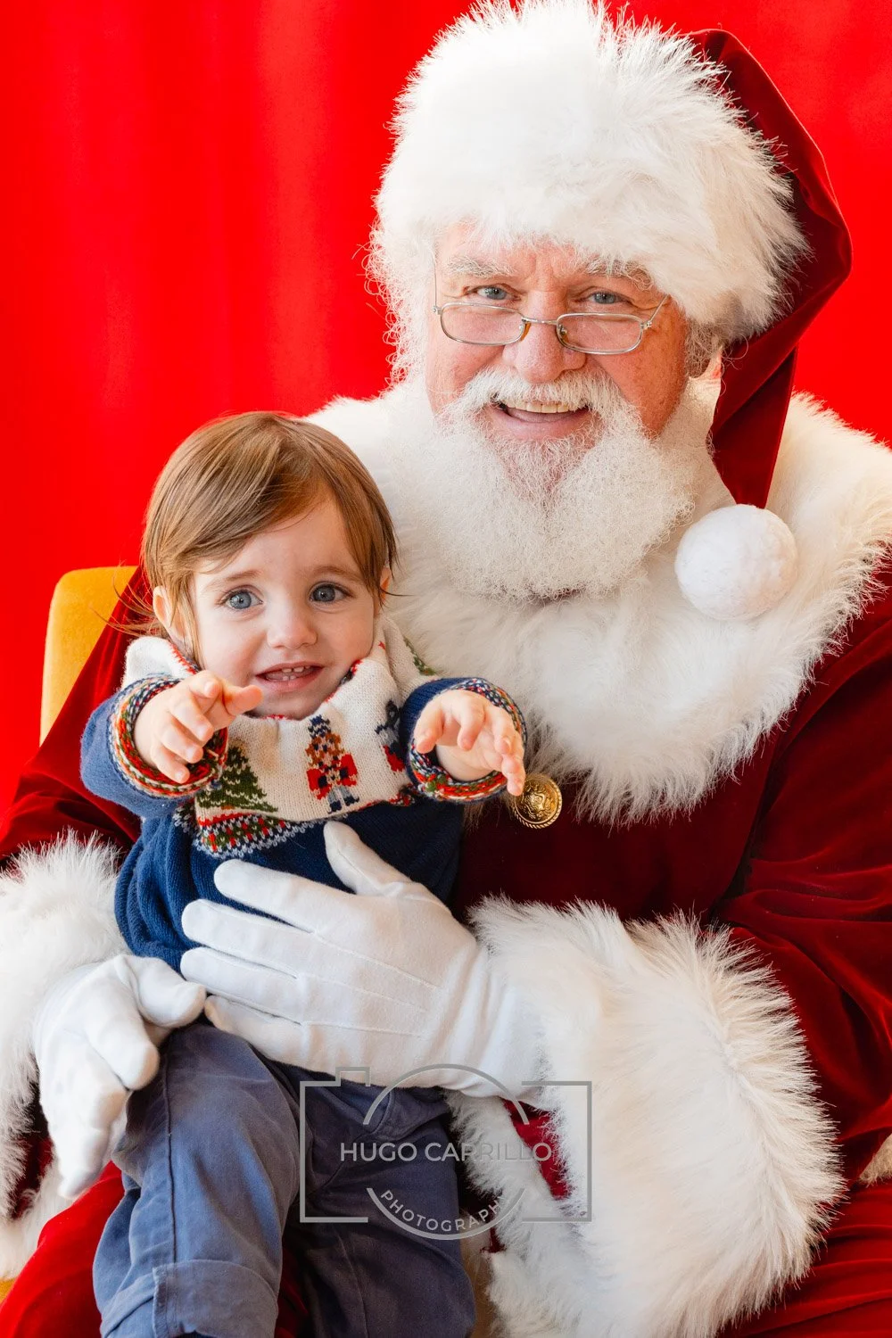 A young boy sitting on Santa Claus' lap, both smiling, with a red background. Santa is dressed in traditional red and white attire, wearing glasses, and the boy is wearing a Christmas sweater.