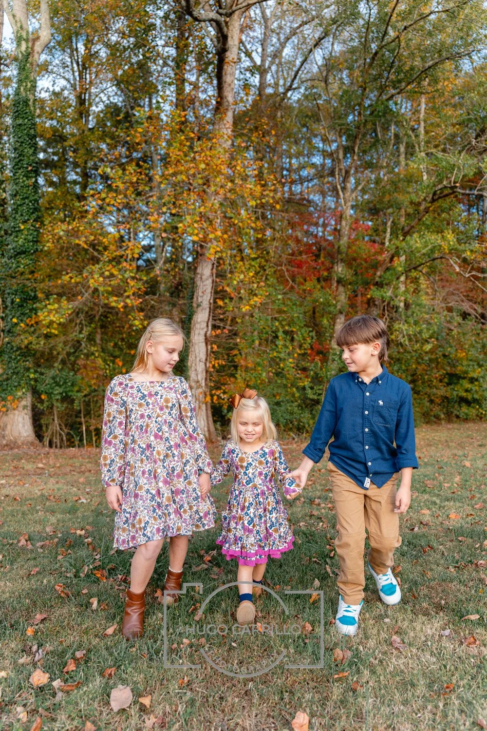 Three children walking hand in hand through a park with autumn trees in the background.