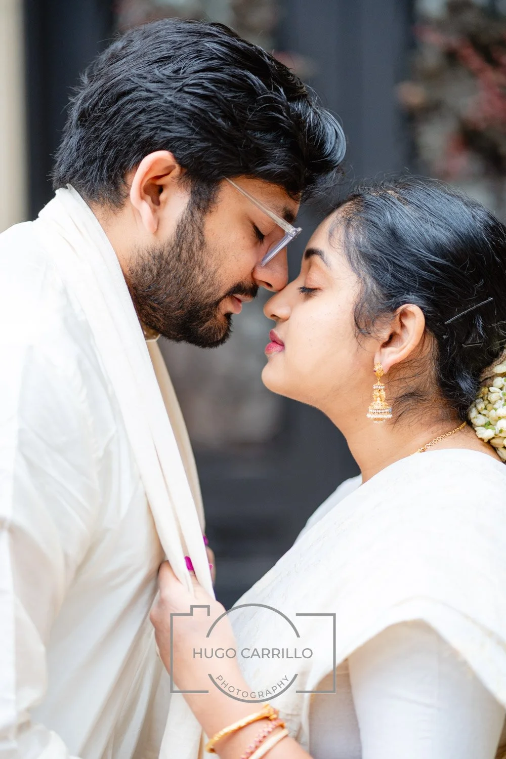 A couple dressed in white traditional Indian attire with their foreheads touching and eyes closed during a romantic moment.
