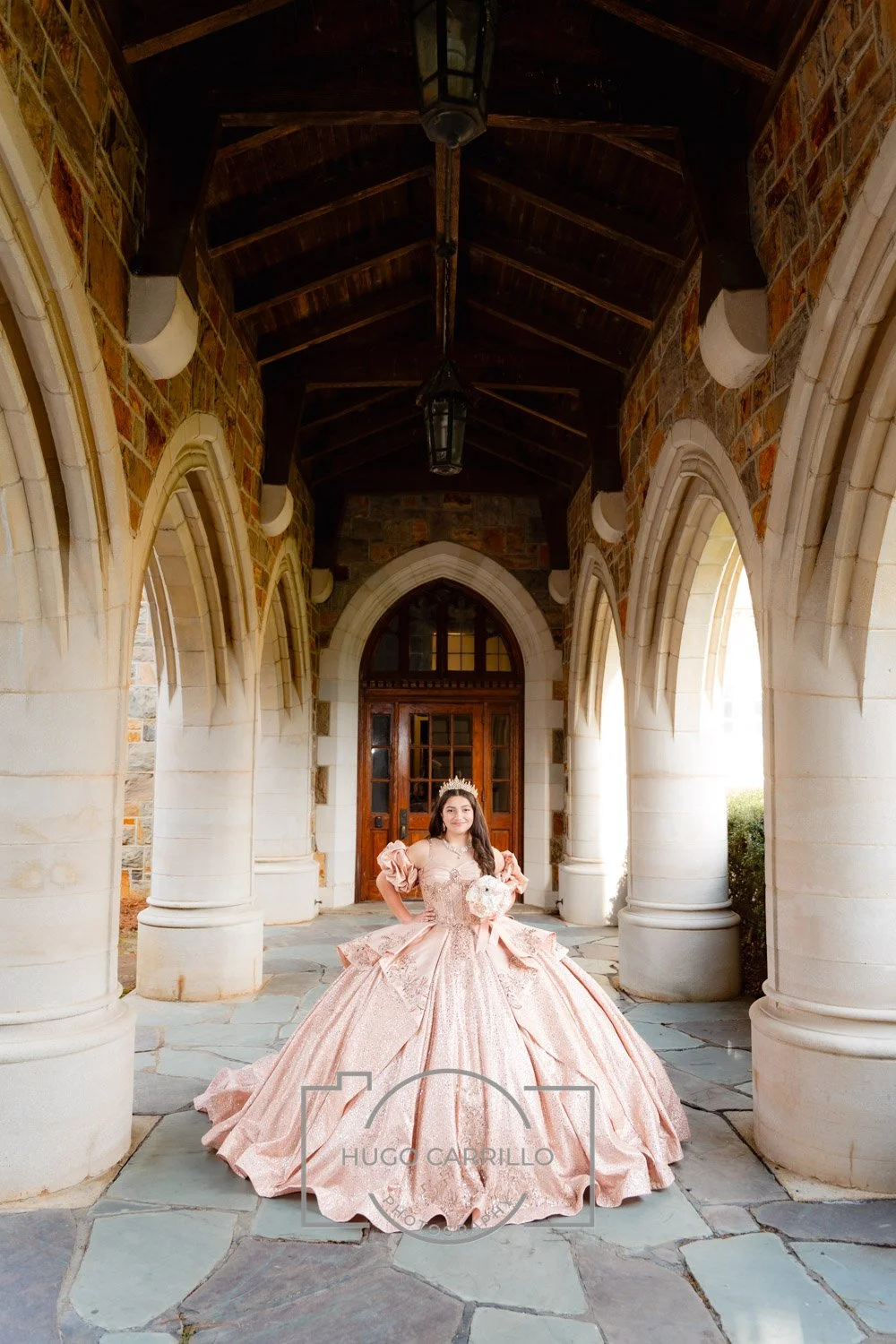 A quinceañera in a light pink fancy ball gown with puffed sleeves, ornate bodice, and wide skirt, wearing a tiara and holding a bouquet, standing beneath a stone archway on a porch of a castle-like building.