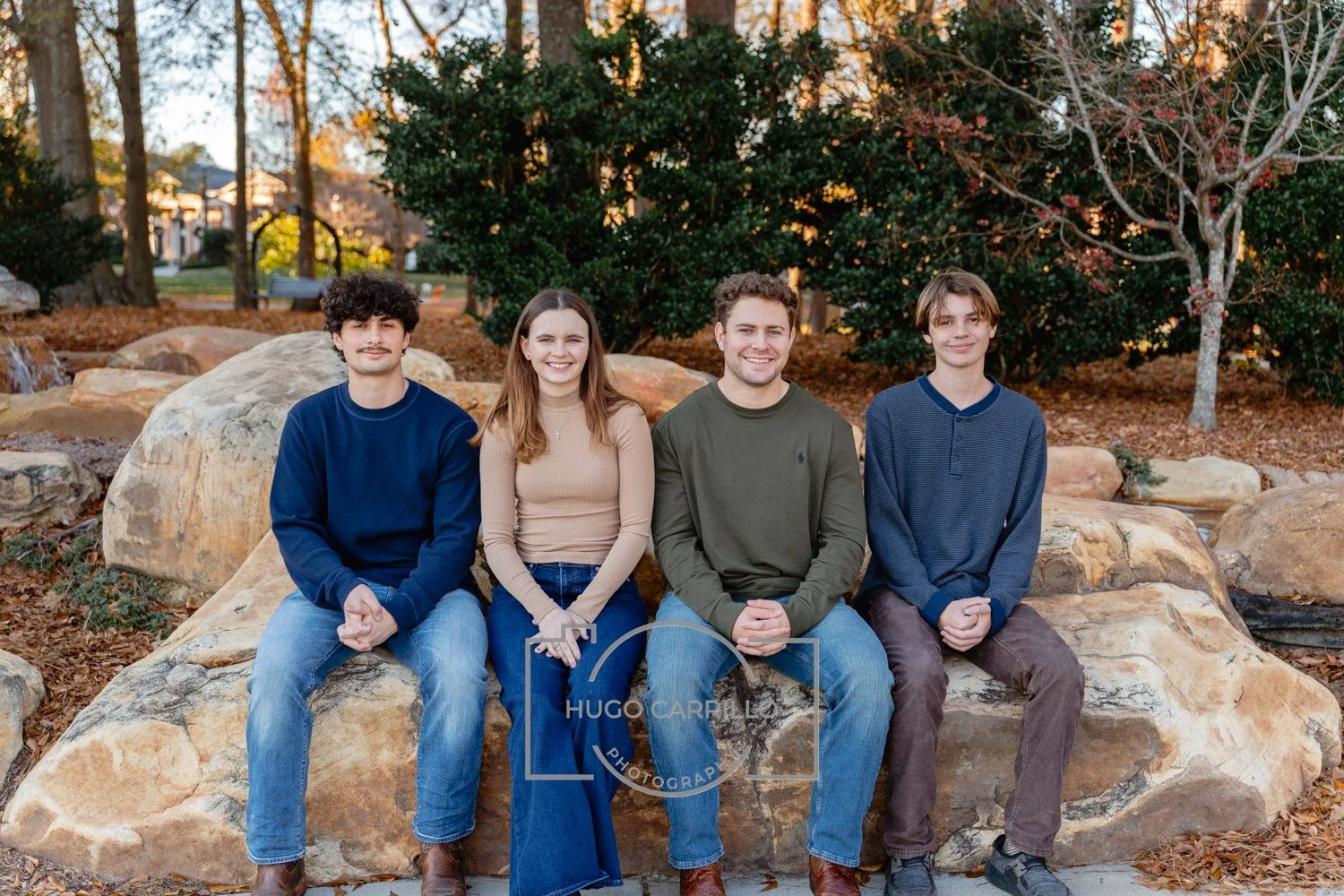 A group of four young people sitting on a large rock outdoors in autumn, smiling at the camera.