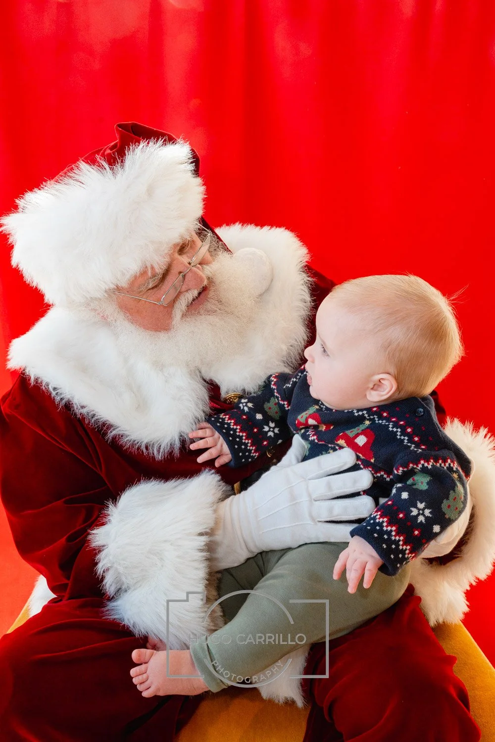Santa Claus in a red suit with white fur trim holding a young child, both looking at each other, with a red backdrop.